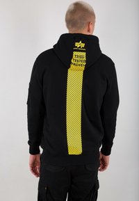 Uomo con capelli corti che indossa una felpa nera con un pannello verticale a strisce gialle e il logo Alpha Industries sulla schiena, in piedi su uno sfondo grigio.