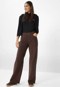 Weite braune Cordhosen mit Taschen, kombiniert mit einem schwarzen, enganliegenden Oberteil und einer schwarzen, strukturierten Weste. Schlichte Schuhe runden das Outfit ab.