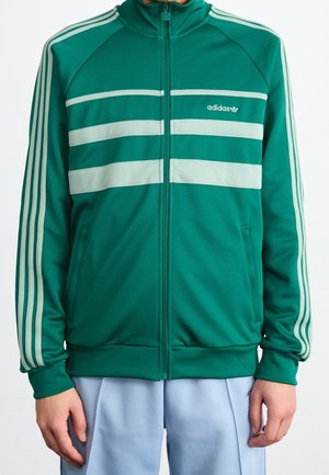 Personne portant une veste de survêtement Adidas verte avec des bandes blanches sur les manches et la poitrine, et un pantalon bleu clair.