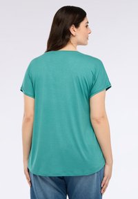 Tyrkysové tričko s krátkým rukávem, uvolněný střih a zakulacený lem. Měkká textura, ležérní design a minimalní švy. Nosené s modrými džíny.