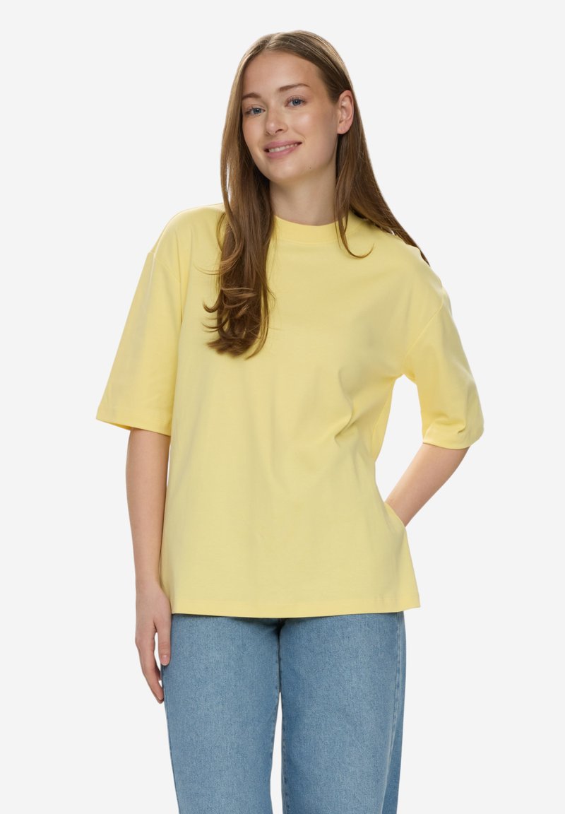 Femme aux longs cheveux bruns souriante, portant un t-shirt jaune oversize et un jean bleu clair, debout avec une main dans le dos.