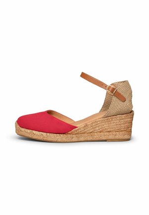 Alpargata de cuña con puntera cerrada roja, talón de yute tejido y correa ajustable para el tobillo en tono tostado con hebilla sobre fondo blanco.
