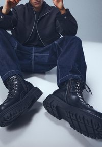 Cizme de luptă negre din piele cu talpă groasă, având ochiuri decorative și șireturi, asortate cu pantaloni din denim albastru închis și o geacă asortată.