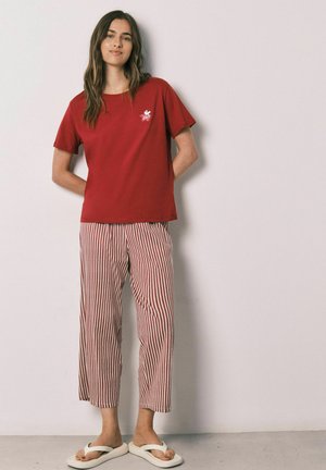 Donna con i capelli lunghi che indossa una maglietta rossa, pantaloni a righe rosse e bianche e infradito bianche, in piedi contro un muro semplice.