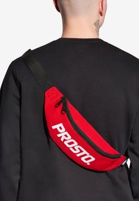 Czerwona saszetka nerkowa o zakrzywionym kształcie, z dużym białym logo i zamkiem błyskawicznym. Nosi się ją na skos przez czarną koszulkę. Regulowany pasek.