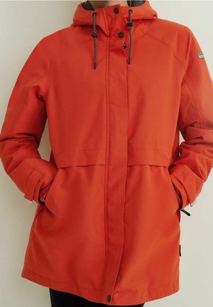 Orange vandtæt jakke med hætte, sorte snore, forreste knapper og lommer, båret af en person med hænderne i lommerne.