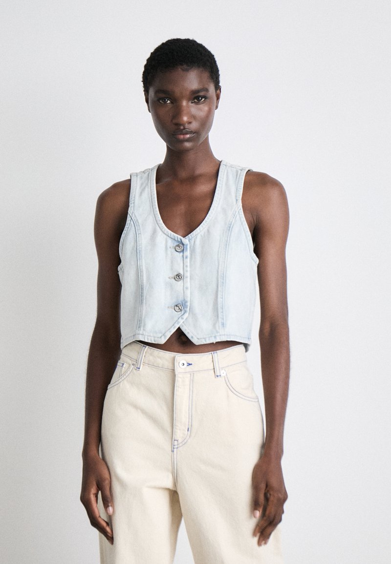 Lyseblå cropped denimvest med dyp V-hals, tre metallknapper og kontrastsøm, kombinert med lyse kremfargede høyt midjepants.