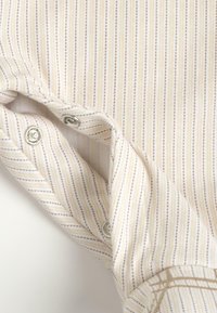 Chemise rayée en crème clair et beige, avec des boutons-pression en argent, en tissu doux et avec des coutures discrètes. Rayures verticales avec des motifs à pois.