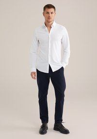 Fehér hosszú ujjú, galléros ing gombokkal, tengerészkék nadrággal és fekete fűzős cipővel párosítva. Egyszerű dizájn, sima anyagtextúra.