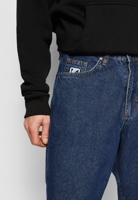 Mørkeblå denimjeans med klassisk snitt. Foran er det synlig søm og et merke på lommen. En svart hettegenser er synlig på armen.