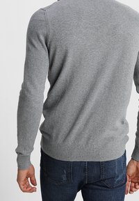 Maglione a maglia grigia con collo rotondo. Presenta maniche lunghe e polsini e orlo a coste. Texture liscia, silhouette aderente. Indossato sopra jeans scuri.