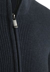 Marineblauer, gerippter Strickpullover mit Reißverschlussdetails. Verfügt über eine glatte Textur und einen metallischen Reißverschluss-Zug. Nahaufnahme des Materials und Designs.