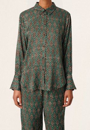 Personne portant une chemise boutonnée à manches longues et un pantalon assorti avec un motif géométrique en losanges verts, marron et blanc.