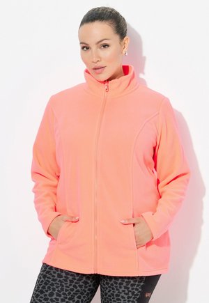 Frau in einer leuchtend korallenfarbenen Fleecejacke mit Reißverschluss, die die Hände in den Taschen hat, und schwarzen gemusterten Leggings vor einem schlichten weißen Hintergrund.