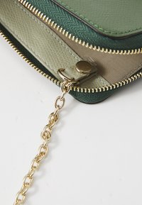 Portafoglio verde e beige con zip, caratterizzato da un materiale testurizzato, accessori in oro e una tracolla con anelli intrecciati.