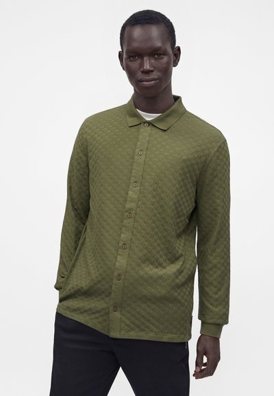Homme portant une chemise verte texturée à manches longues boutonnée sur un sous-t-shirt blanc, debout devant un fond clair uni.