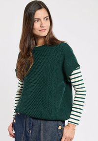 Vrouw met lang bruin haar, gekleed in een donkergroene kabelgebreide mouwloze trui over een wit en donkergroen gestreept langemouwen shirt en een spijkerrok.