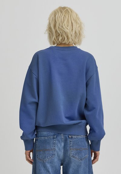 Sweat-shirt bleu à coupe décontractée, col rond, épaules tombantes, poignets et ourlet côtelés. Stylisé avec un jean large bleu clair.