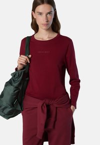 Camisa de manga larga burdeos con el logo "NORTH SAILS", combinada con pantalones a juego atados a la cintura y un bolso verde de hombro.