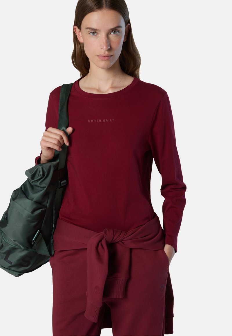 Camisa de manga larga burdeos con el logo "NORTH SAILS", combinada con pantalones a juego atados a la cintura y un bolso verde de hombro.