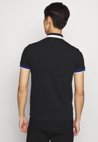 En person sedd bakifrån bär en svart kortärmad polo med vit och blå kant på ärmarna och kragen mot en enfärgad bakgrund.