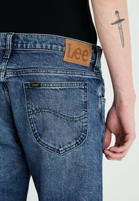 Sinimärkiset denim-farkut, joissa on nahkainen Lee-brändilogo takana, kahdella taskulla ja ommelkoristeilla. Haalistunut pinta ja rento istuvuus.