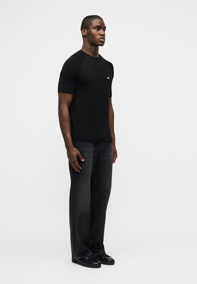 Άνδρας που στέκεται φορώντας μαύρο κοντομάνικο πουκάμισο με λογότυπο, σκούρο γκρι τζιν και μαύρα παπούτσια σε απλό γκρι φόντο.