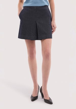 Frau trägt einen dunkelgrauen Plisseerock und schwarze High Heels, steht mit den Händen in den Taschen vor einem schlichten Hintergrund.