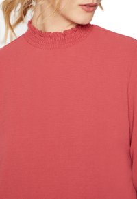 Punainen paita rypytetyllä korkealla kaula-aukolla, pehmeästä materiaalista ja pitkillä hihoilla, korostaa yksinkertaista ja hienostunutta muotoilua.