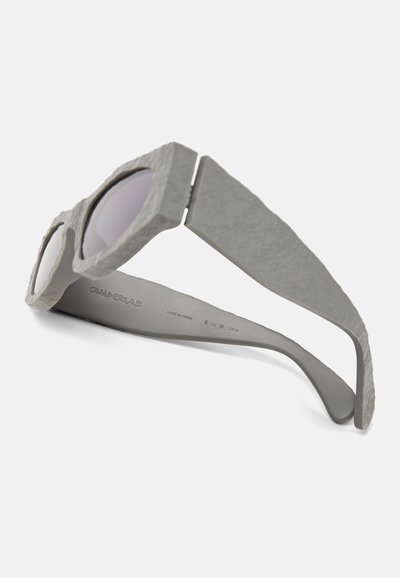 Grijze zonnebril heeft een gestructureerd frame met ronde lenzen en dikke pootjes. De binnenkant van de pootjes is glad en toont merken en productdetails.