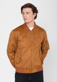 Junger Mann trägt eine braune Wildleder-Bomberjacke mit Fronttaschen und ein weißes Unterhemd, blickt zur Seite vor einem schlichten Hintergrund.