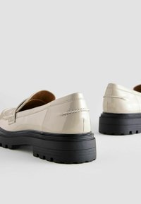Beige patent leather loafers with a thick black platform sole, featuring a decorative strap and stitching details on the back.