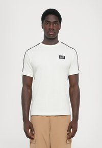 White cotton t-shirt with black accents on shoulders, fitted design, round neckline, and a small branded patch on the chest.