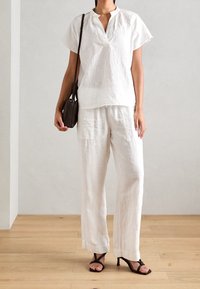 Haut en lin blanc à manches courtes avec un décolleté en V, assorti à un pantalon ample en lin blanc. Porte avec des sandales à talons marron et un petit sac sombre.