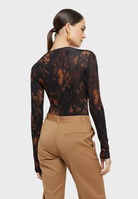 Dugi rukav majica u crnoj boji s smeškastim uzorkom, glatke teksture. Uparen s bež hlačama s prugicama koje imaju bočne džepove.