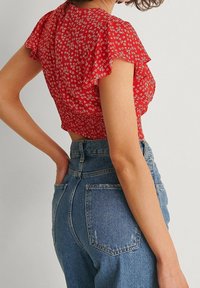Top corto floreale rosso con maniche a volantino corte e schiena arricciata, abbinato a jeans in denim blu a vita alta con tasche posteriori visibili.