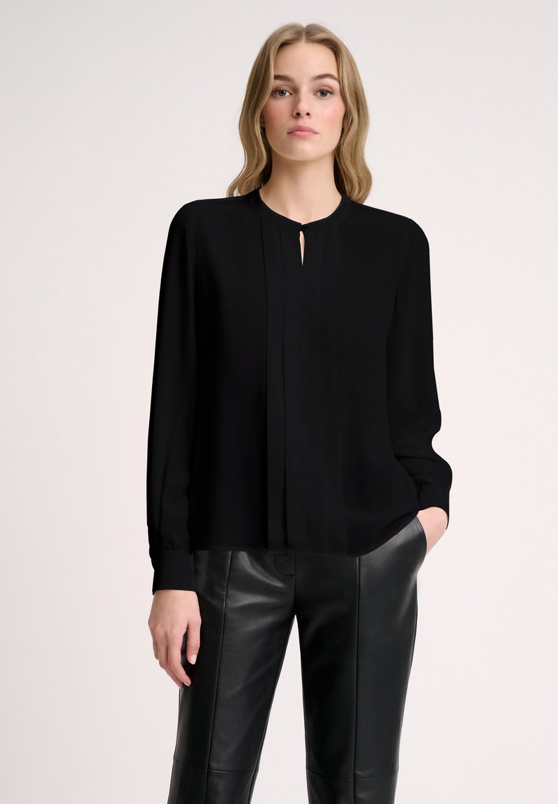 Blusa nera a maniche lunghe con scollo rotondo, dettaglio a chiave e design plissettato. Abbinata a pantaloni di pelle neri. Tessuto liscio e leggero.
