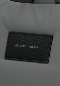 Grau Tasche mit einem strukturierten schwarzen Etikett, auf dem das "TOM TAILOR" Logo in dezentem grauen Schriftzug integriert ist.
