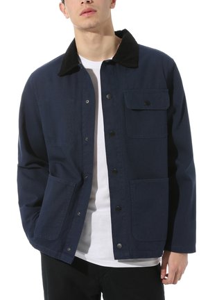 Jeune homme portant une veste boutonnée bleu marine avec un col noir, une poche à rabat sur le devant et deux poches plaquées inférieures, par-dessus une chemise blanche.