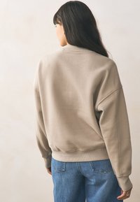 Beżowa bluza o luźnym kroju, z prążkowanymi ściągaczami na rękawach i u dołu. Noszona z niebieskimi dżinsami. Gładka faktura, minimalne detale i prosty design.