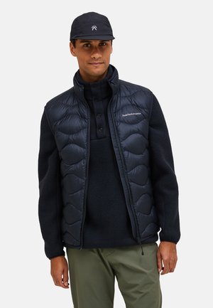 Vesta puf neagră cu un design matlasat, asociată cu un top din fleece închis la culoare și o sapcă întunecată. Vesta are un închidere cu fermoar și detalii cu logo.