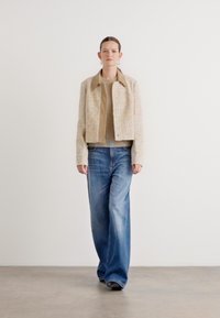 Beige Tweedjacke mit Kragen, Knopfleiste und strukturiertem Muster, kombiniert mit hellblauen weit geschnittenen Jeans. Schlichte Schuhe runden den Look ab.