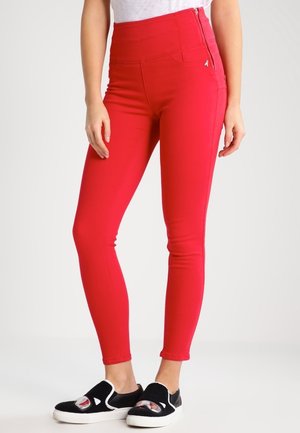 Kobieta nosząca czerwone, obcisłe spodnie z wysokim stanem i bocznym suwakiem oraz czarne wsuwane buty z wzorem postaci z kreskówki na białych podeszwach.