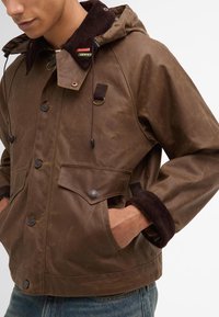 Brown waxed jacket with a corduroy collar, featuring snap buttons, drawstring hood, and large front pockets. Textured, water-resistant finish.