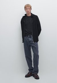 Schwarze Reißverschluss-Hoodie über einem schwarzen T-Shirt, kombiniert mit dunklen Denim-Jeans und braunen Schnürschuhen. Einfacher, lässiger Stil.