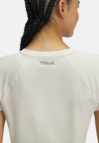 Viegls, elpojošs sporta krekls krēmkrāsā, ar apaļu kaklu, perforētu tekstūru un smalku "TRAIL" logo aizmugurē.