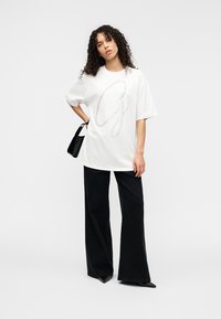 Λευκό μπλουζάκι με μεγάλο γκρι γραφικό γράμμα «G», συνδυασμένο με μαύρο παντελόνι φαρδύ και μυτερά μαύρα τακούνια. Η μοντέλο κρατάει μια μικρή μαύρη τσάντα.