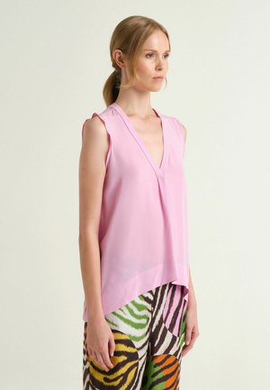 Donna con capelli biondi raccolti in una coda di cavallo, che indossa un top senza maniche color rosa chiaro con scollo a V e pantaloni colorati a righe zebrate, vista di lato.