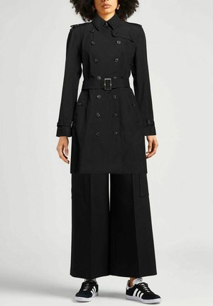 Donna che indossa un trench doppio petto nero con cintura, pantaloni neri a gamba larga e sneakers Adidas nere e bianche.