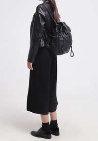 Sac à dos en cuir noir, forme arrondie, fermeture à cordon, et bretelles réglables. Porté avec une veste en cuir noire et un large pantalon noir.
